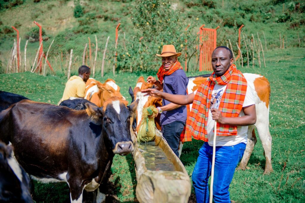Diary farming A group of people gathered outdoors near green vegetation, one person wearing a yellow hat and orange outfit, another in a red checkered vest and blue pants, with a woven basket visible."
