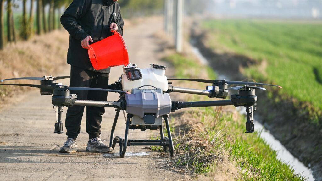 A person fills a large agricultural drone with liquid from a red container beside a farm field.